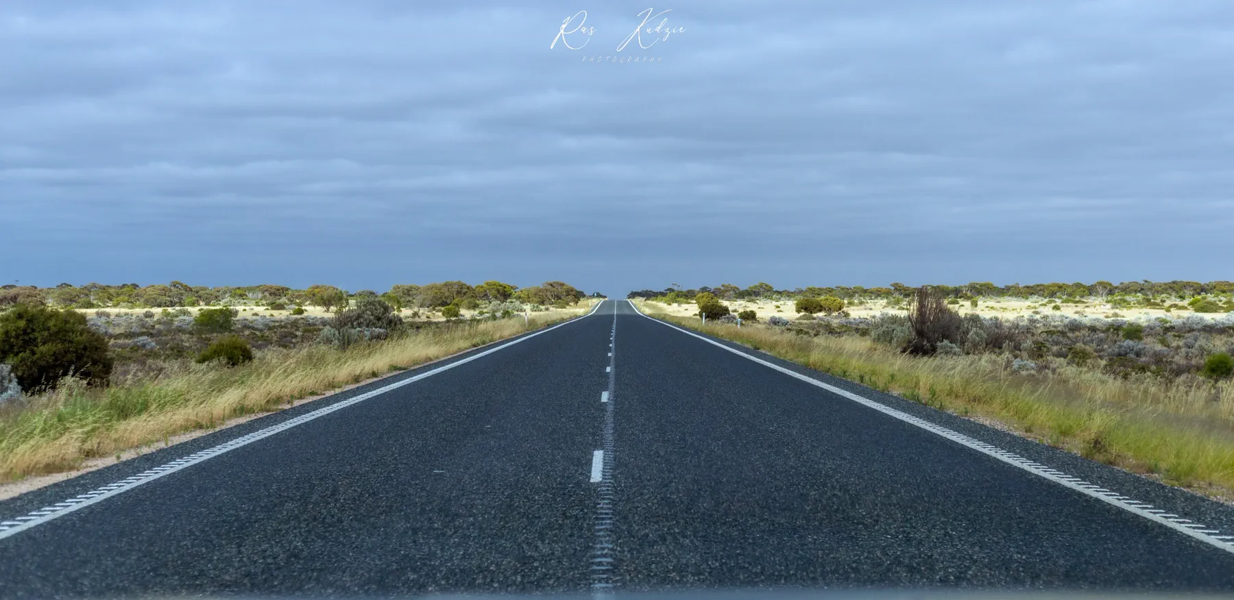 A long open road running through Western Australian scrub beneath a cloudy sky