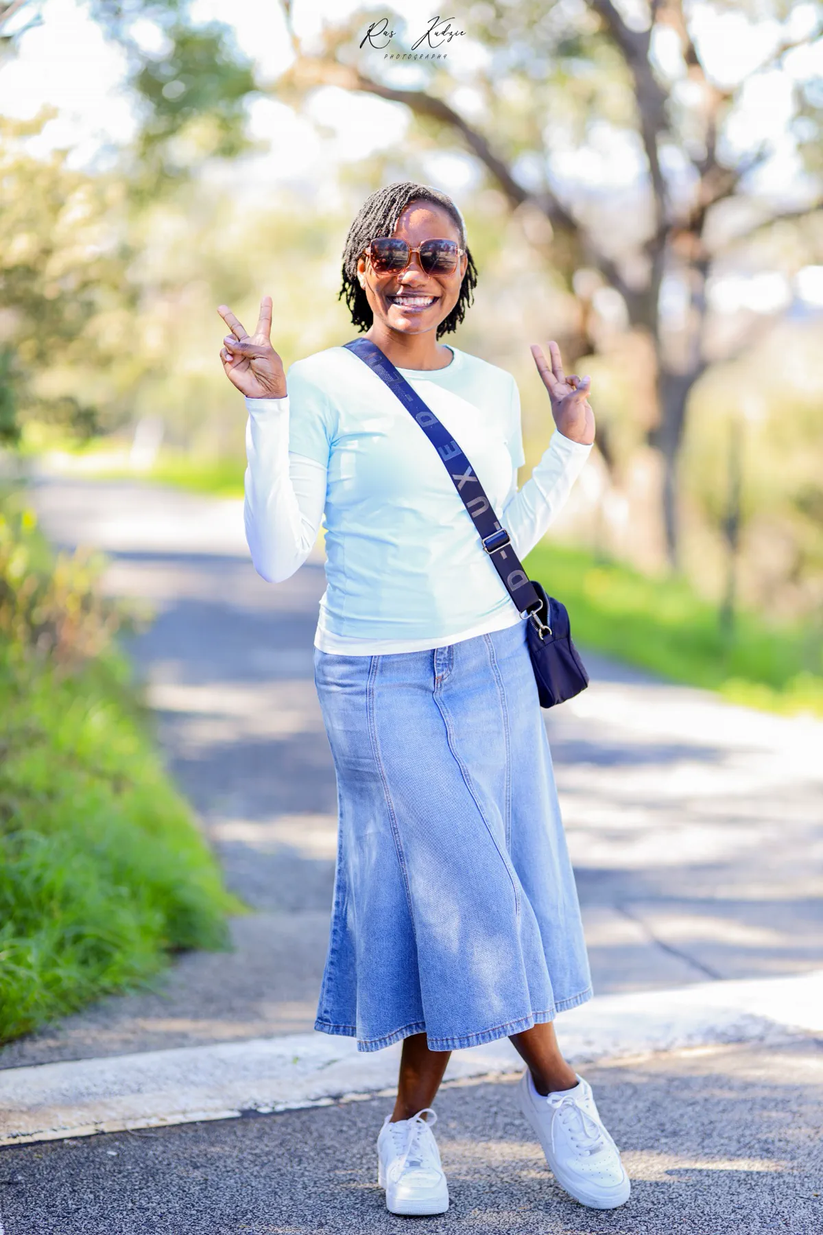 A woman smiling and raising two peace signs on a sunlit path